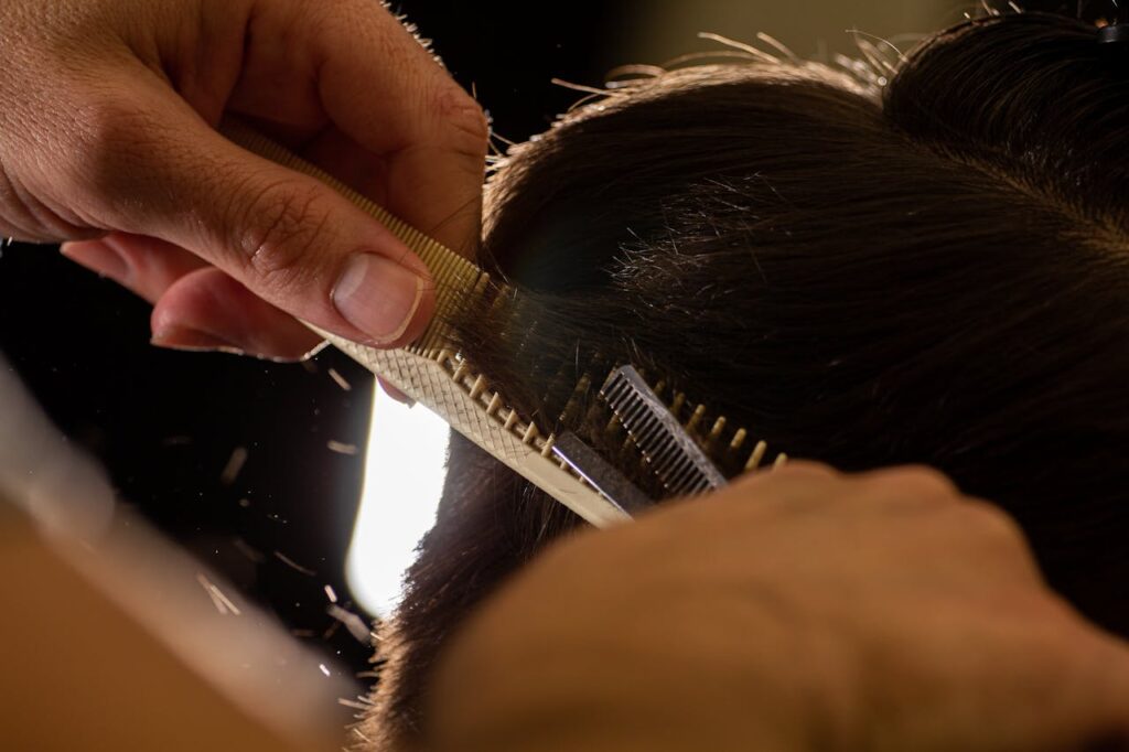 pexels-photo-6487888 Close-up of a stylist trimming hair with a comb and scissors indoors.